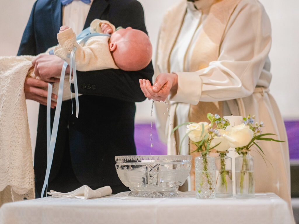 Vauva miehen sylissä, pappi valelemassa vettä vauvan päähän kasteessa.
