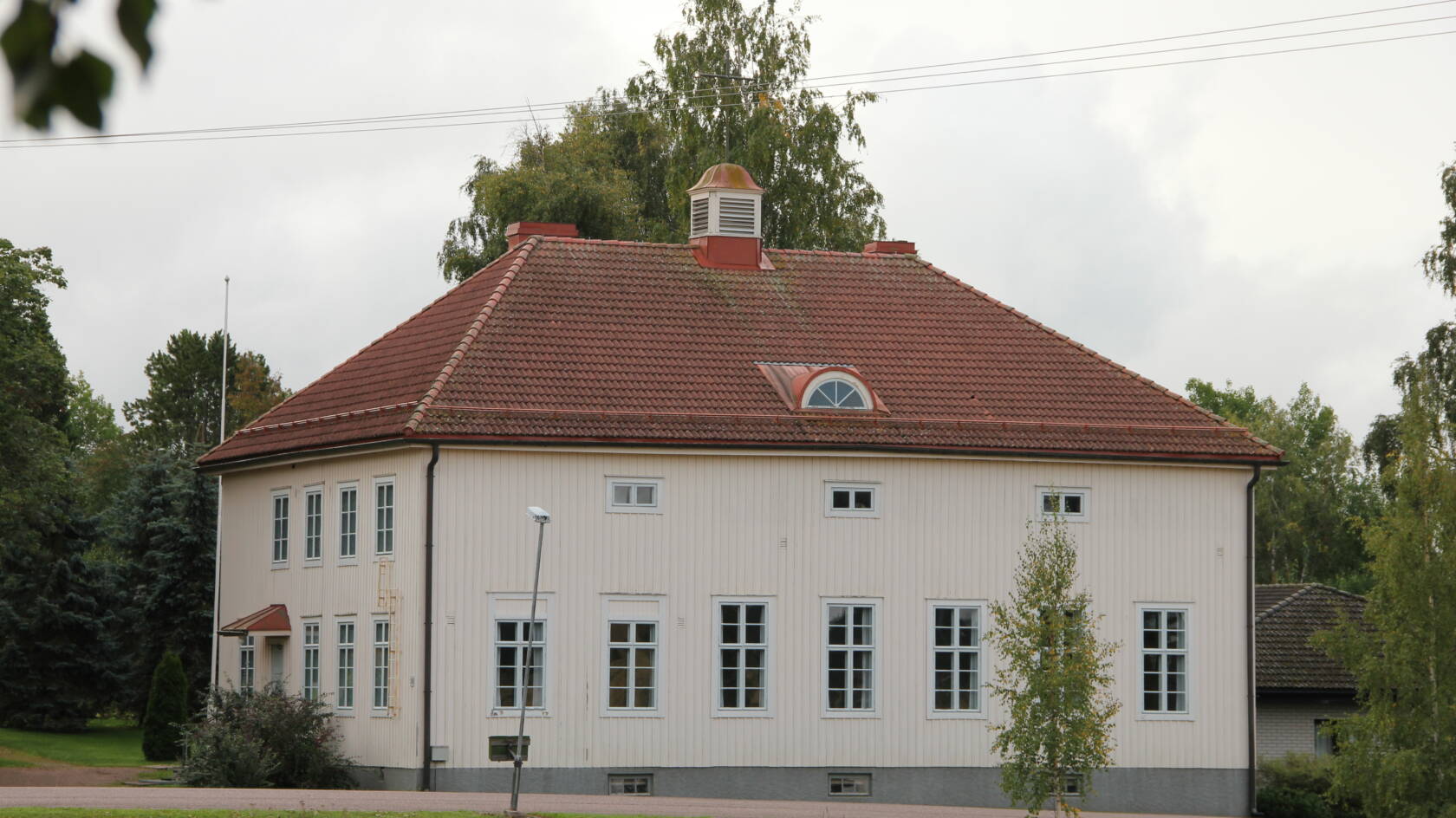 Vaalea lautaverhottu kaksikerroksinen puurakennus punaisella tiilikatolla. 1930-luvulta. Kapeat korkeat ikkunat.