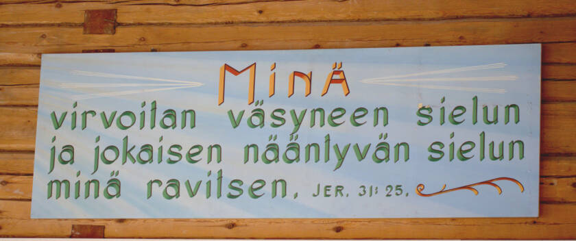 Raamatunlausetaulu seurakuntatalon salin yläosassa. Taustalla hirsiseinä. Jer 31:25.