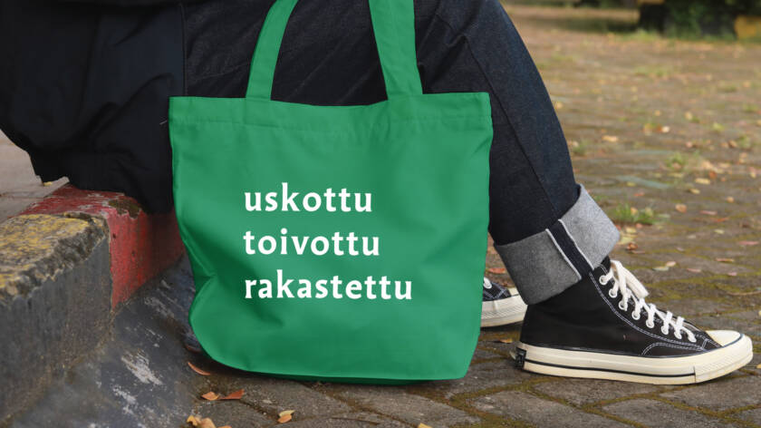 Ihminen, jonka jalat näkyvät, istuu katukivetyksen reunalla vieressään kassi, jossa infoa seurakuntavaaleista.