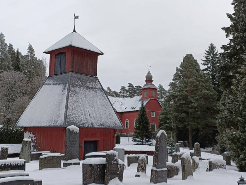 Vanhan hautausmaan hautakiviä ja kellotorni taustalla puukirkko talvisessa maisemassa.
