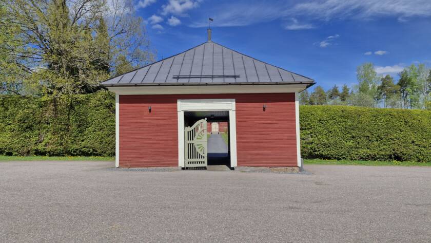 Puuporttirakennus, jonka läpi kuljetaan hautausmaa, kummallakin puolella korkea kuusiaita.