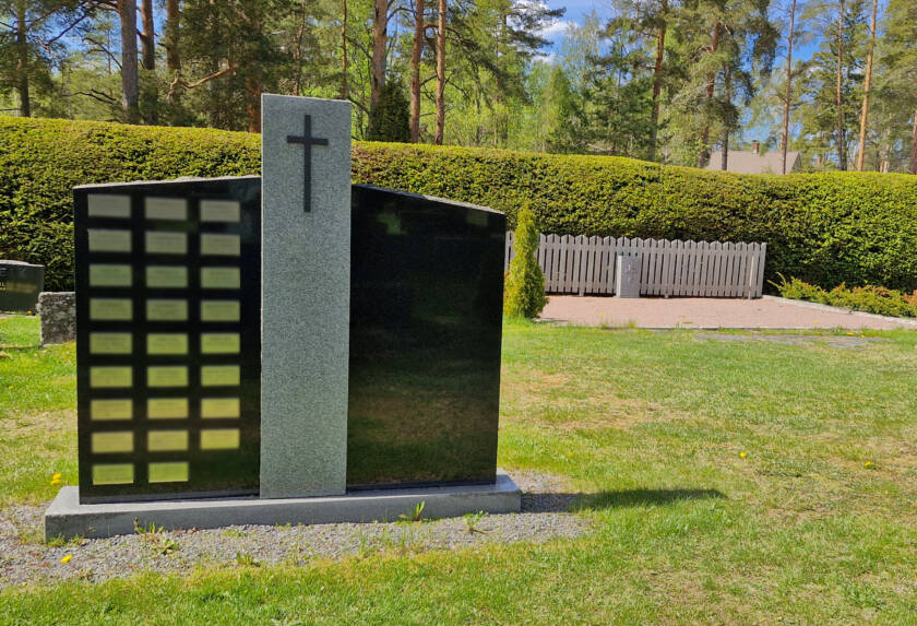 Iso kivipaasi, jossa nimilaattoja, taustalla hiekkainen uurna-alue.