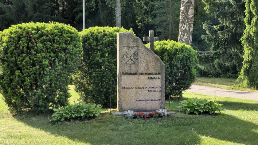 Kivipaasi, jonka viistossa yläkulmassa risti, taustalla pensaita.