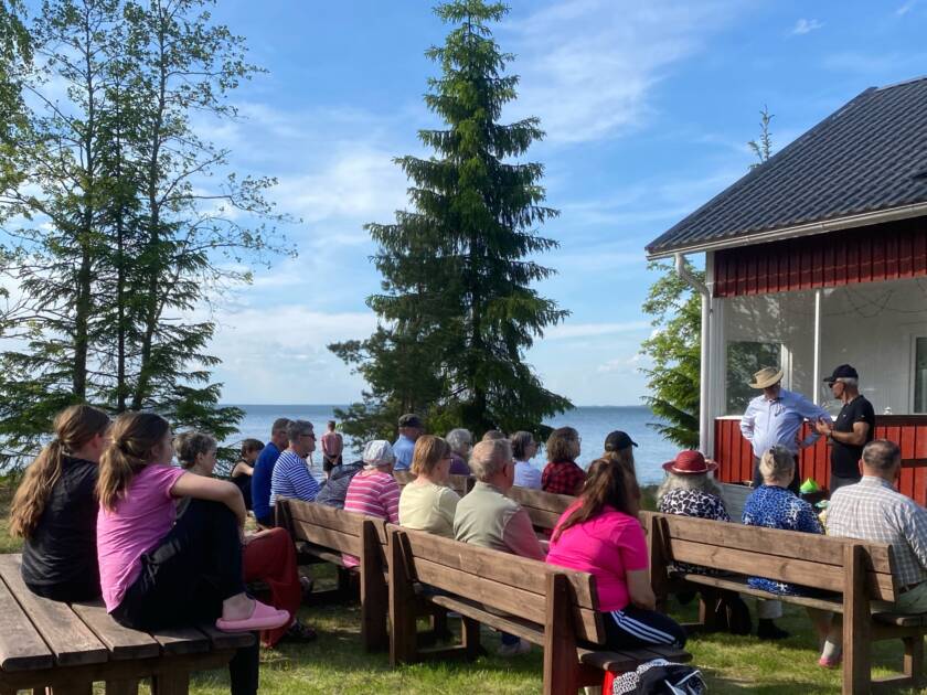 Ihmisiä kuuntelemassa vierasta aurinkoisena kesäisenä iltana mökkirannassa.