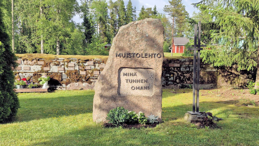 Korkea kivipaasi, jonka reunat rosoiset, taustalla kiviaita, jossa muistolaattoja.