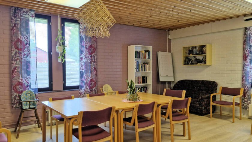 Kokoushuoneen keskellä pöytä ja tuolit, taustalla pieni sohva ja värikkäät verhot.