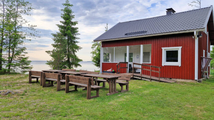 Pieni mökki järven rannalla taustalla järvi ja nurmikolla pöytiä ja penkkejä.