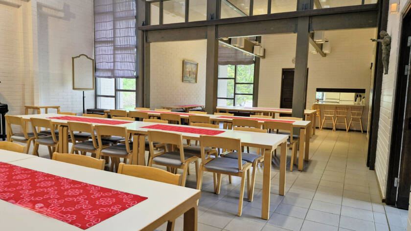 Pieni seurakuntasali tuoleineen ja pöytineen taustalla tarjoilupöytätila.