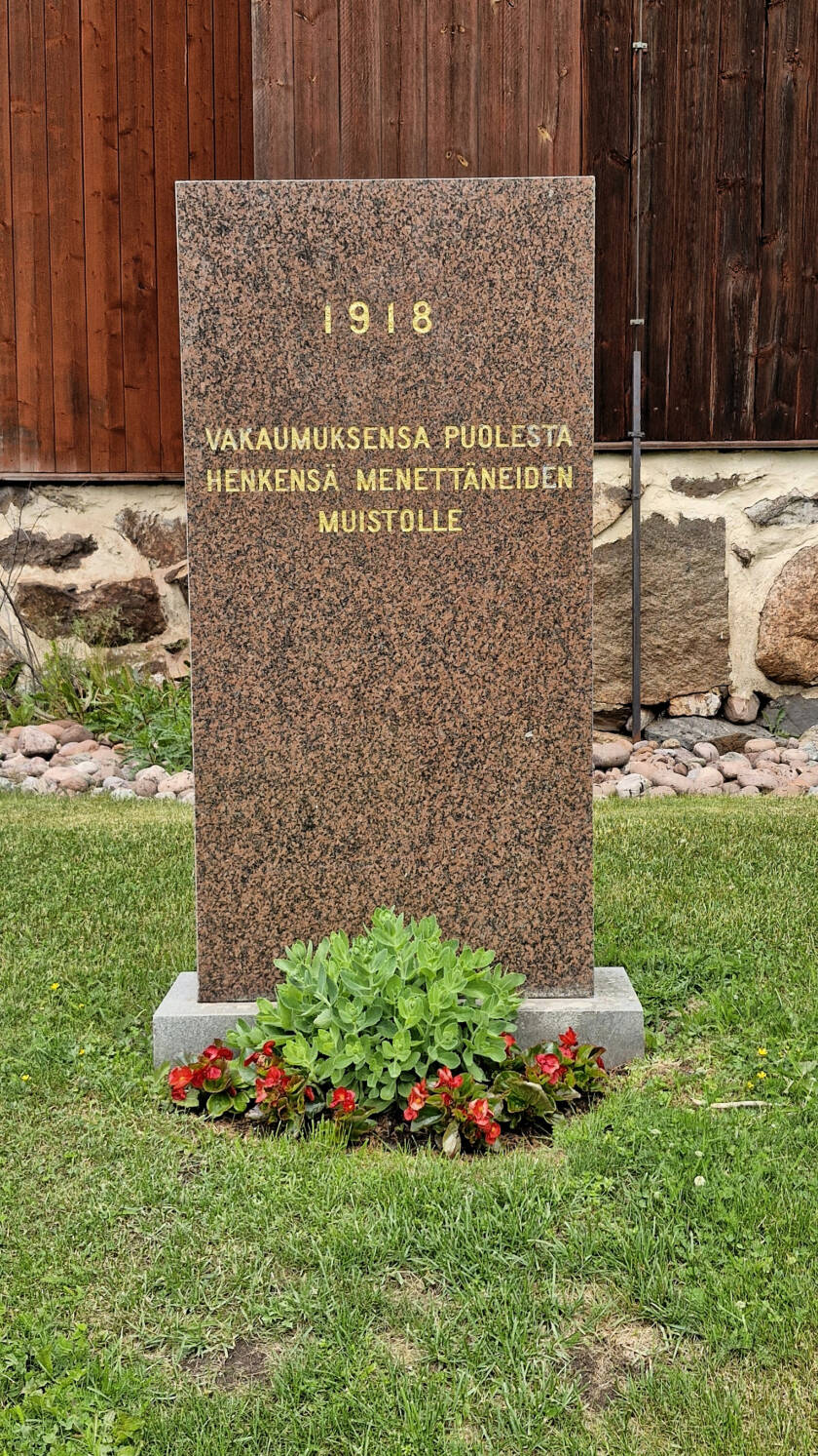 Kivipaasi kirkon seinän lähellä sekä kukkia muistomerkin edessä.
