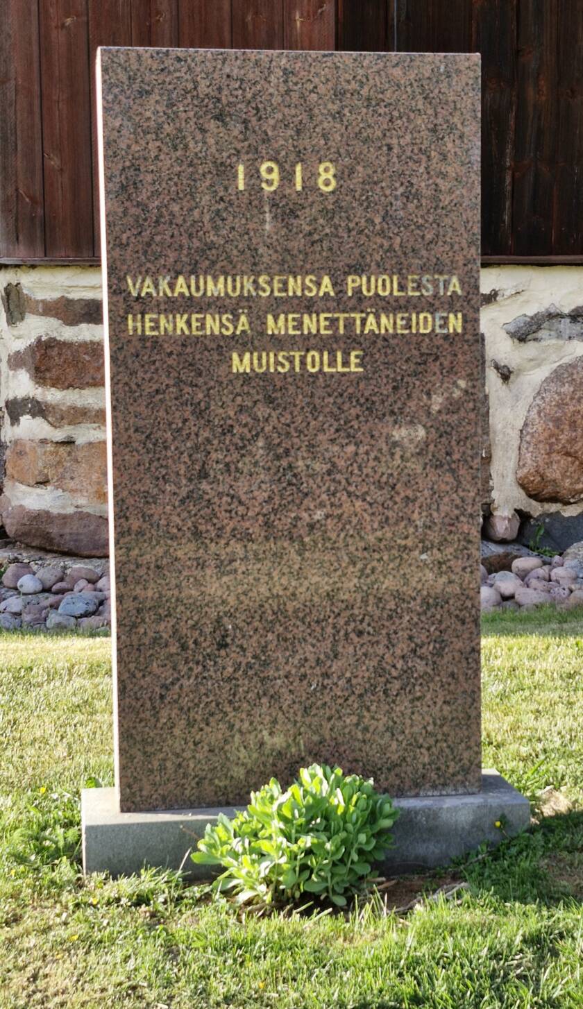 Kivipaasi kirkon seinän lähellä sekä kukkia muistomerkin edessä.