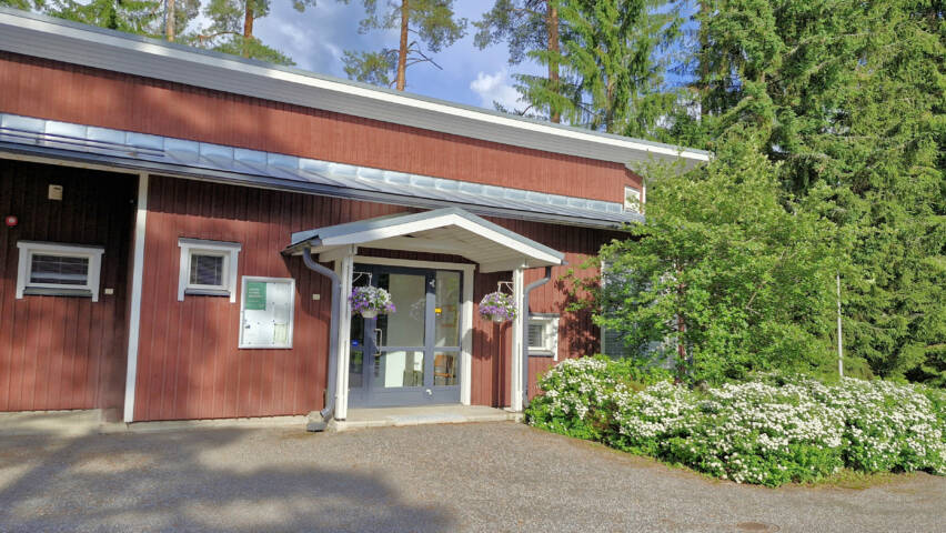 Puinen matala rakennus, joka edessä kukkivia pensaita ja korkean pensasmainen puu.